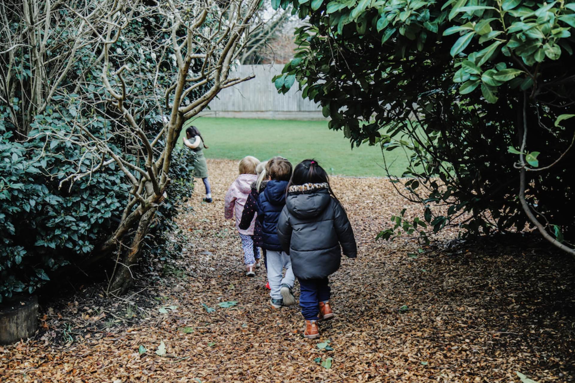Exploring outdoors at Kindred Bromley North Nursery and Pre-school A group of children are walking single-file along a leafy path in a garden at the nursery.